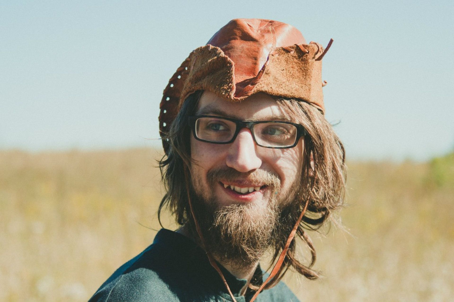 Man smiling outdoors wearing glasses and a weathered leather cowboy hat.